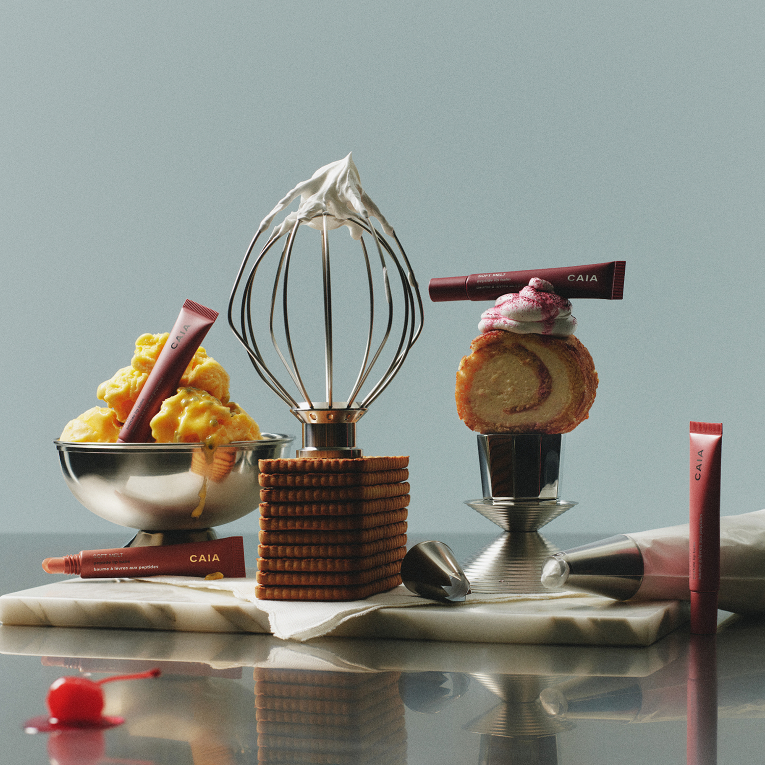 Still life of CAIA Soft melt peptide lip balm products styled with desserts and baking tools—whipped cream on a whisk, cookies, a pastry, and a piping bag—set on a marble surface with a soft, minimal background.